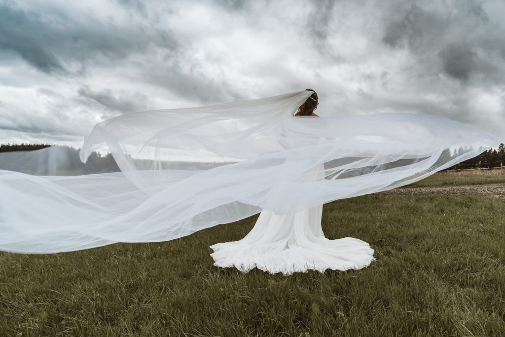 Schleierfoto, Braut steht auf einer Wiese und der Schleier weht im Wind