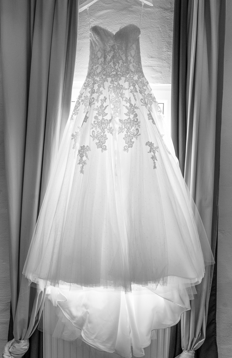 Brautkleid auf dem Kleiderbügel am Fenster. Licht scheint durch das Brautkleid. Links und rechts sind Vorhänge zu sehen