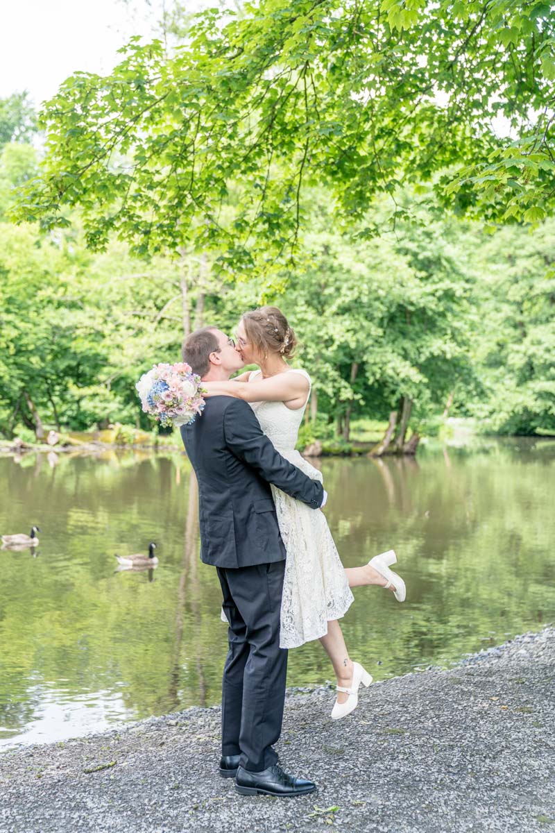 Brautpaar am Weiher, er hält Sie etwas hoch und sie streckt ein Fuß nach hinten. Dabei umarmt Sie ihn mit dem Brautstrauß und beide küssen sich.