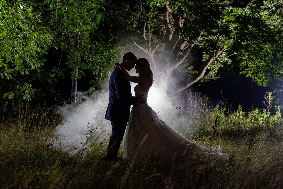 Brautpaar um dunklen. Beide umarmen sich. Von hinten Blitzlicht und Nebel. Das ganze vor einem Baum