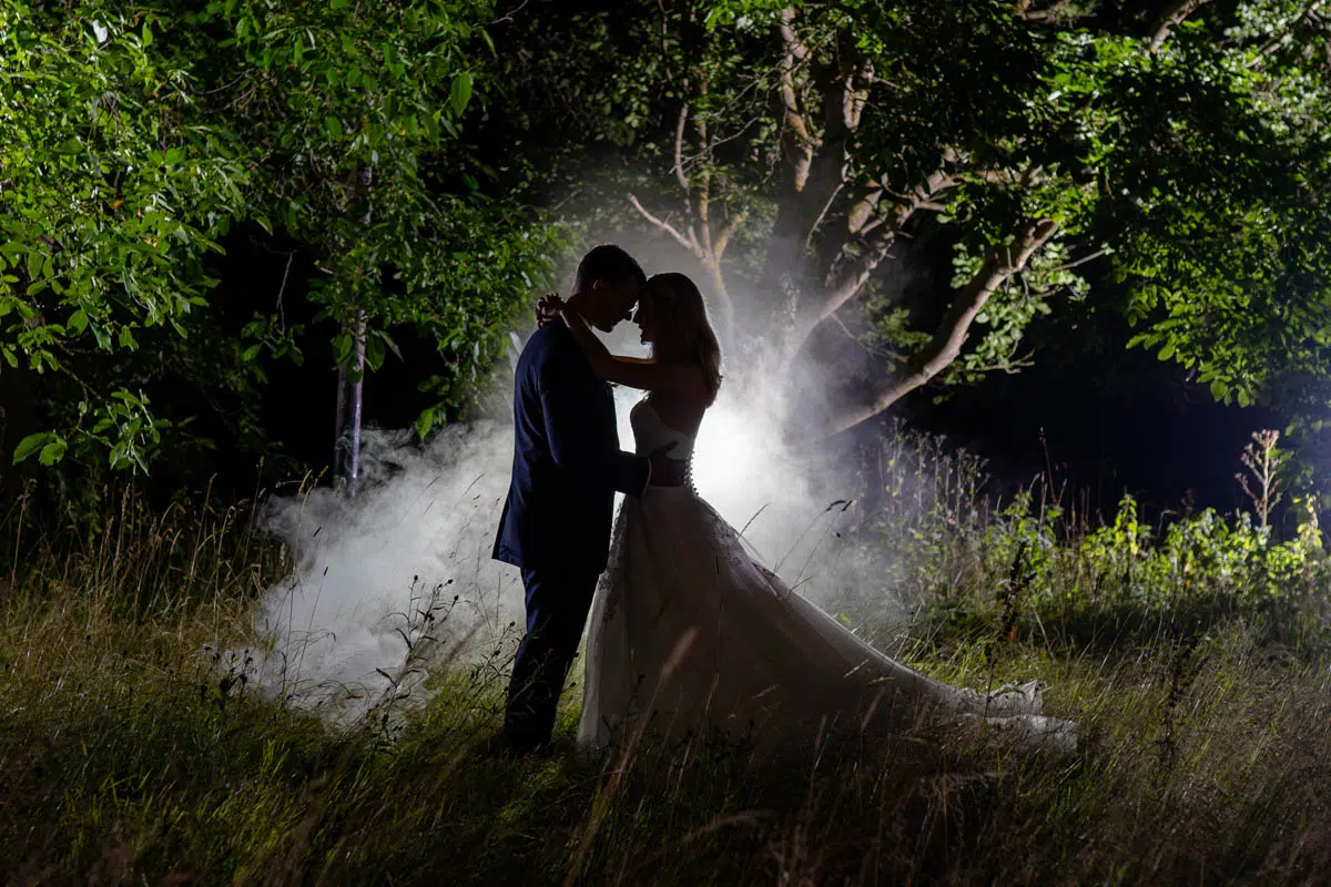 Brautpaar um dunklen. Beide umarmen sich. Von hinten Blitzlicht und Nebel. Das ganze vor einem Baum